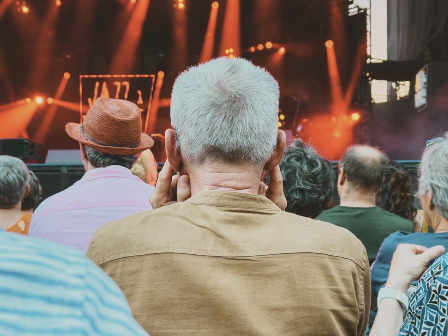 An old man plugging his ears with his fingers at a concert, seen from the back.